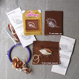 May include: A Jiffy Stitchery embroidery kit featuring a spider conch shell design. The kit includes brown embroidery floss, a brown fabric square, and instructions. The kit is packaged in a clear plastic bag with a gold and white label.