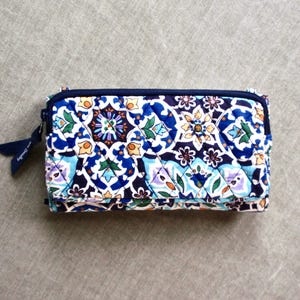 May include: A quilted wallet with a vibrant floral pattern in shades of blue, white, and purple. The wallet has a navy blue zipper and a matching fabric pull tab. The overall design is intricate and colorful.