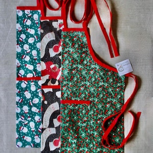 May include: Three Christmas aprons with red trim and ties. One apron has a green background with white snowmen, another has a black background with red and white designs, and the third has a green background with red berries.