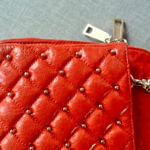 Pu&ograve; includere: Primo piano di un portafoglio in pelle trapuntata rossa con borchie color argento. Il portafoglio presenta una cerniera argentata e una tracolla a catena argentata. La pelle ha un motivo a rombi.