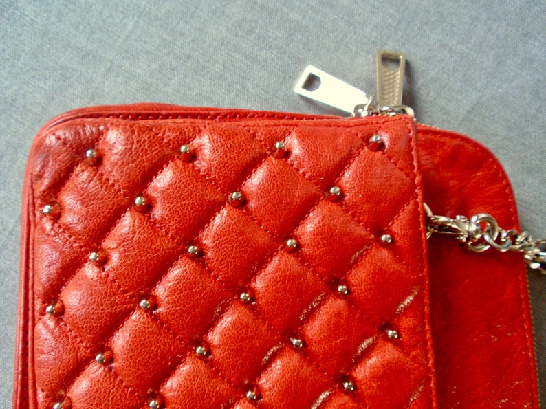 Pu&ograve; includere: Primo piano di un portafoglio in pelle trapuntata rossa con borchie color argento. Il portafoglio presenta una cerniera argentata e una tracolla a catena argentata. La pelle ha un motivo a rombi.