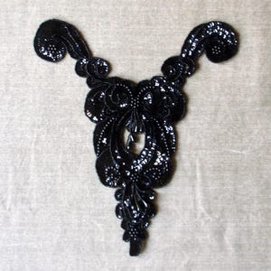 May include: A black sequined applique with an ornate, symmetrical design. The applique features a central oval shape with a teardrop pendant, flanked by swirling, scroll-like elements. The surface is covered in shiny sequins.