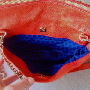 Pu&ograve; includere: Borsa a mano in pelle arancione aperta con cerniera dorata e tracolla a catena. L'interno &egrave; foderato in tessuto blu con un motivo a pois neri. La borsa &egrave; aperta, rivelando l'interno.