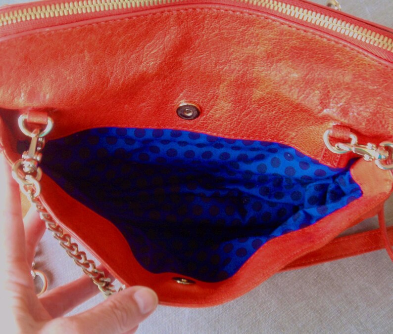 Pu&ograve; includere: Borsa a mano in pelle arancione aperta con cerniera dorata e tracolla a catena. L'interno &egrave; foderato in tessuto blu con un motivo a pois neri. La borsa &egrave; aperta, rivelando l'interno.