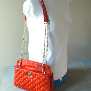 Pu&ograve; includere: Una borsa trapuntata, rosso brillante, con tracolla a catena e finiture color oro. La borsa ha una forma rettangolare con chiusura a patta e cerniera. La borsa &egrave; esposta su un manichino bianco.