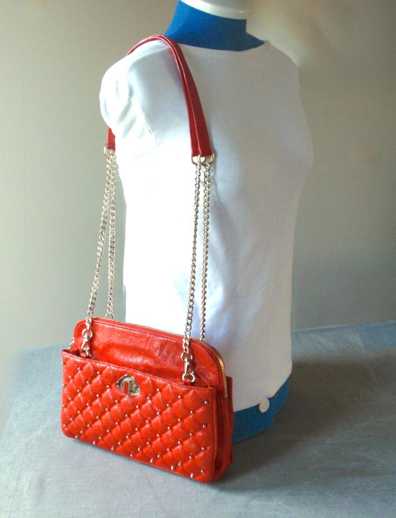 Pu&ograve; includere: Una borsa trapuntata, rosso brillante, con tracolla a catena e finiture color oro. La borsa ha una forma rettangolare con chiusura a patta e cerniera. La borsa &egrave; esposta su un manichino bianco.