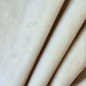 May include: Close-up of a soft, beige fabric with a smooth, velvety texture. The fabric is folded in a way that creates a series of gentle curves and folds.