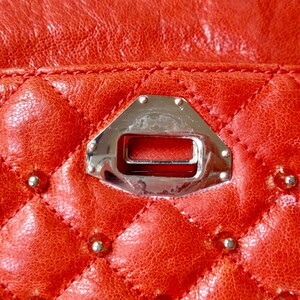 Pu&ograve; includere: Primo piano di una borsa in pelle trapuntata rossa. La pelle presenta un motivo a rombi con piccole borchie argentate alle intersezioni. Una chiusura in metallo argentato &egrave; centrata, con un'apertura rettangolare e bordi rivettati.
