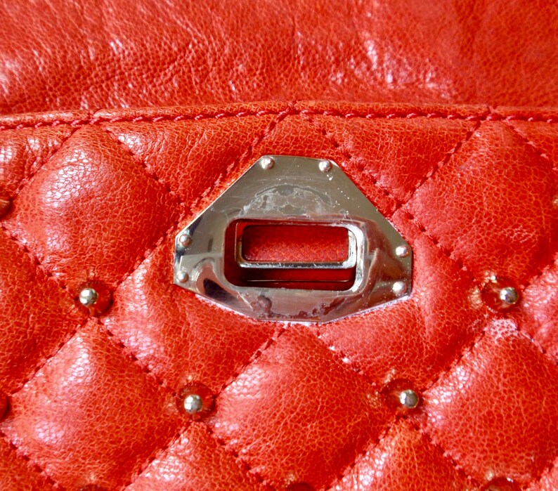 Pu&ograve; includere: Primo piano di una borsa in pelle trapuntata rossa. La pelle presenta un motivo a rombi con piccole borchie argentate alle intersezioni. Una chiusura in metallo argentato &egrave; centrata, con un'apertura rettangolare e bordi rivettati.