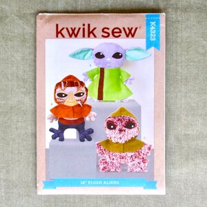 May include: A sewing pattern titled "kwik sew" featuring three plush alien designs. The aliens are in green, orange, and pink, with the text "18" PLUSH ALIENS" at the bottom. The pattern is on a light brown background.