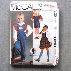 May include: Vintage McCall's sewing pattern featuring children's clothing designs. The pattern includes illustrations of a young girl in a blue jumpsuit, a romper, and a plaid jumper. The pattern is labeled "8718" and "Child Size 5".