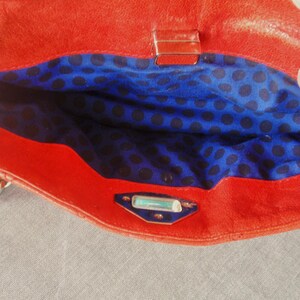 Pu&ograve; includere: Borsa a mano in pelle rossa aperta, che rivela un interno blu a pois neri. La borsa ha una tracolla a catena e una chiusura argentata. L'interno &egrave; visibile.