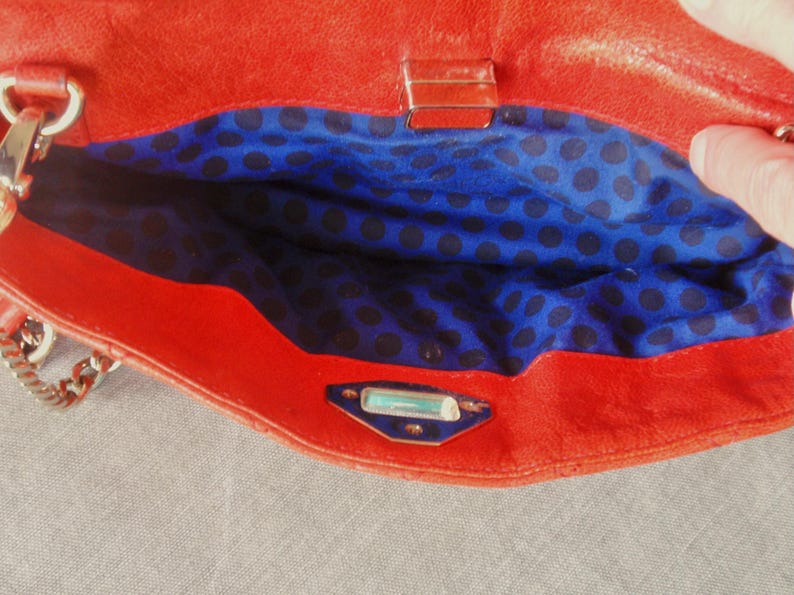 Pu&ograve; includere: Borsa a mano in pelle rossa aperta, che rivela un interno blu a pois neri. La borsa ha una tracolla a catena e una chiusura argentata. L'interno &egrave; visibile.