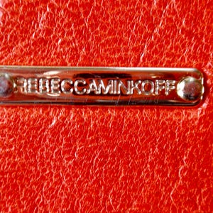 Pu&ograve; includere: Primo piano di una superficie in pelle rossa brillante e strutturata. Una placca metallica rettangolare color oro &egrave; fissata alla pelle, con la scritta in rilievo "REBECCAMINKOFF". La placca &egrave; fissata con due piccoli rivetti arrotondati.