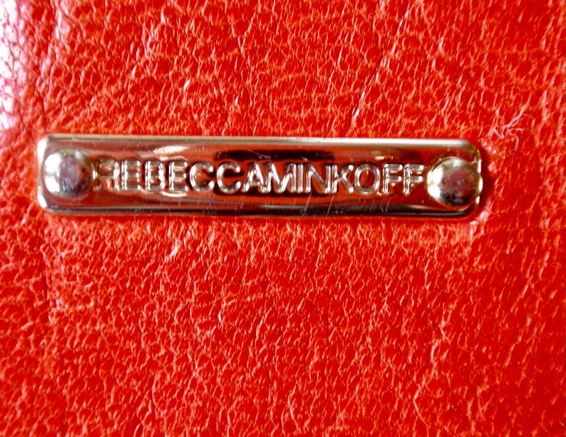 Pu&ograve; includere: Primo piano di una superficie in pelle rossa brillante e strutturata. Una placca metallica rettangolare color oro &egrave; fissata alla pelle, con la scritta in rilievo "REBECCAMINKOFF". La placca &egrave; fissata con due piccoli rivetti arrotondati.