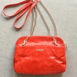 Pu&ograve; includere: Una borsa a tracolla in pelle rosso vivo con una tracolla a catena dorata e un manico in pelle rossa abbinato. La borsa ha una forma rettangolare con chiusura a zip e una piccola placca metallica dorata sul davanti.