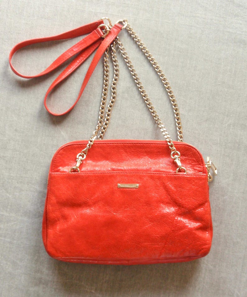 Pu&ograve; includere: Una borsa a tracolla in pelle rosso vivo con una tracolla a catena dorata e un manico in pelle rossa abbinato. La borsa ha una forma rettangolare con chiusura a zip e una piccola placca metallica dorata sul davanti.