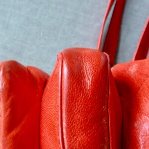 Pu&ograve; includere: Primo piano di una borsa in pelle rosso vivo. La borsa ha una superficie strutturata e cuciture visibili. Anche i cinturini sono rossi e sembrano essere fatti dello stesso materiale. La borsa ha un'estetica vintage.