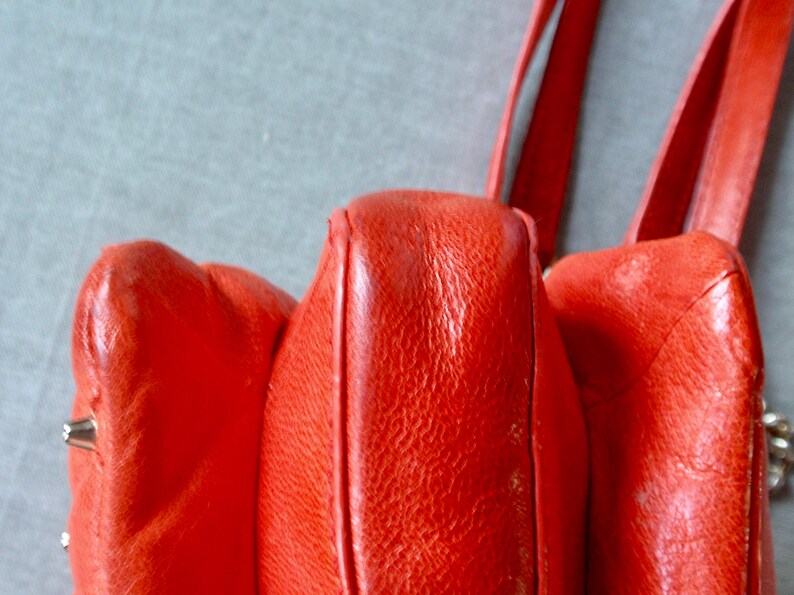 Pu&ograve; includere: Primo piano di una borsa in pelle rosso vivo. La borsa ha una superficie strutturata e cuciture visibili. Anche i cinturini sono rossi e sembrano essere fatti dello stesso materiale. La borsa ha un'estetica vintage.