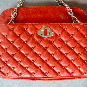 Pu&ograve; includere: Borsa a mano in pelle rossa trapuntata con motivo a rombi e borchie argentate. Ha una tracolla a catena argentata e una chiusura argentata. La patta superiore &egrave; di colore rosso pieno.