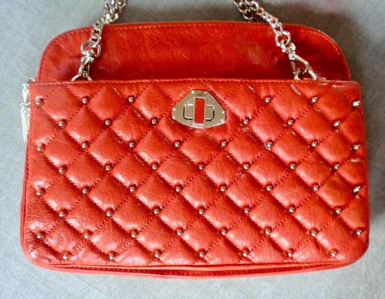 Pu&ograve; includere: Borsa a mano in pelle rossa trapuntata con motivo a rombi e borchie argentate. Ha una tracolla a catena argentata e una chiusura argentata. La patta superiore &egrave; di colore rosso pieno.