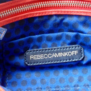 Pu&ograve; includere: Vista interna di una borsa con fodera blu a pois. Un'etichetta blu navy con il marchio "REBECCAMINKOFF" &egrave; cucita all'interno. La borsa ha una cerniera e finiture rosse.
