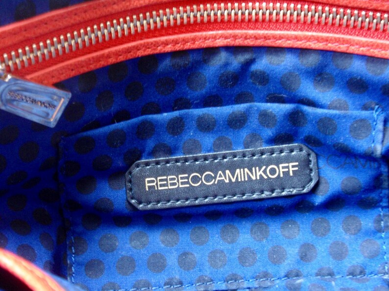 Pu&ograve; includere: Vista interna di una borsa con fodera blu a pois. Un'etichetta blu navy con il marchio "REBECCAMINKOFF" &egrave; cucita all'interno. La borsa ha una cerniera e finiture rosse.
