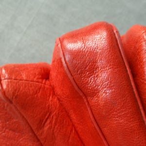 Pu&ograve; includere: Primo piano di una borsa in pelle rosso vivo. La borsa presenta un design trapuntato con cuciture visibili e una superficie strutturata. Una tracolla a catena argentata &egrave; parzialmente visibile.