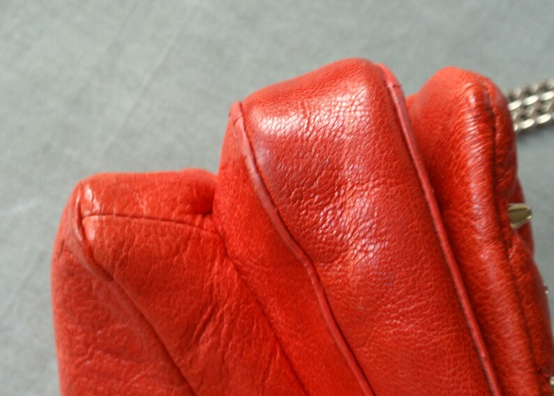 Pu&ograve; includere: Primo piano di una borsa in pelle rosso vivo. La borsa presenta un design trapuntato con cuciture visibili e una superficie strutturata. Una tracolla a catena argentata &egrave; parzialmente visibile.