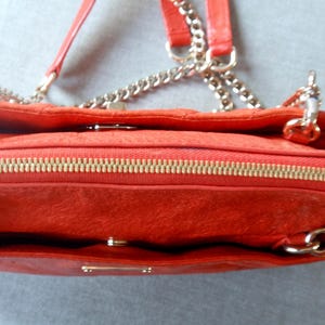 Pu&ograve; includere: Primo piano di una borsa rossa con cerniera dorata e tracolla a catena. La borsa ha una superficie strutturata e una chiusura con patta. La tracolla &egrave; color argento.