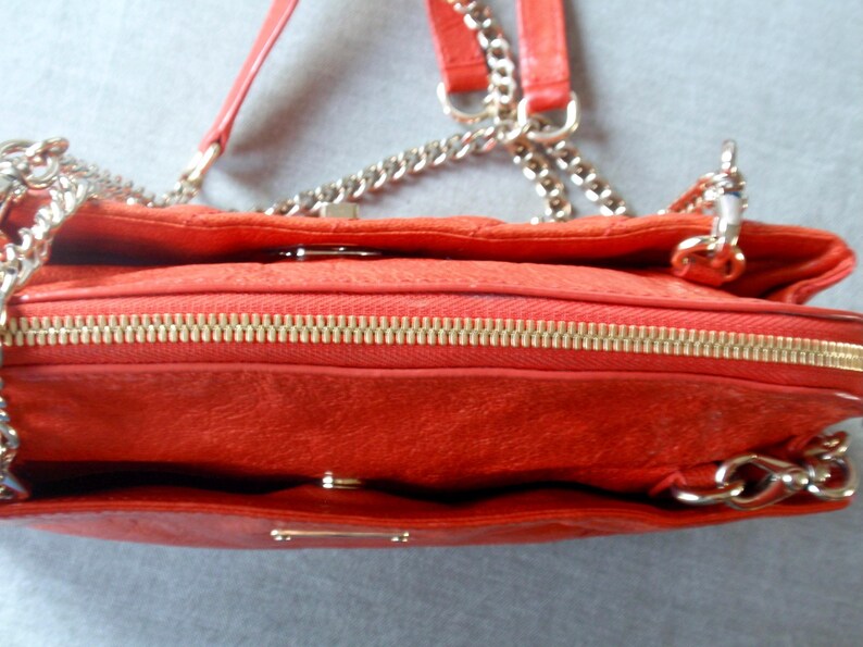 Pu&ograve; includere: Primo piano di una borsa rossa con cerniera dorata e tracolla a catena. La borsa ha una superficie strutturata e una chiusura con patta. La tracolla &egrave; color argento.