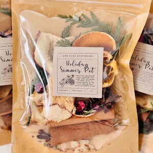 May include: A cellophane bag containing dried ingredients for a simmer pot, including orange slices, cinnamon sticks, and star anise. The label reads "Holiday Simmer Pot" with instructions. The bag is sealed and ready to use.