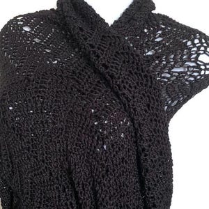 May include: A black, open-weave crochet top. The top features a draped neckline and short sleeves. The crochet pattern creates a delicate, airy texture. The garment is displayed on a white background.