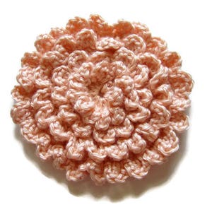 May include: A handmade, peach-colored crochet flower with a detailed, textured design. The flower is round, with multiple layers of petals, and is set against a white background. The craft item is suitable for various DIY projects.