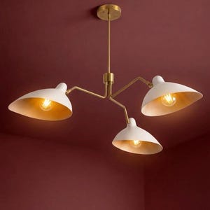 Op de afbeelding: Een moderne plafondlamp met drie witte, bladvormige kappen en goudkleurige interieurs. De armatuur heeft een messingkleurige steel en armen en is gemonteerd op een bordeauxrood plafond.