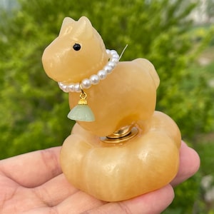 May include: A light orange horse figurine with a pearl necklace and a green pendant. The horse is perched on a round base. The figurine is held in a hand against a blurred green background.