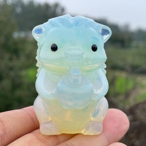 Puede incluir: Una figura de erizo translúcida, azul claro y amarillo, con ojos negros. El erizo sostiene un objeto pequeño. La figura está hecha de un material pulido, posiblemente cristalino.