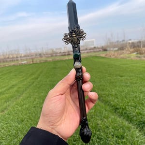 Op de afbeelding: Een zwarte toverstaf met een kristallen punt, versierd met een gouden bij-bedel en groene en grijze stenen. De toverstaf wordt in een hand gehouden tegen een groene gras achtergrond.