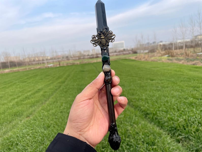 Op de afbeelding: Een zwarte toverstaf met een kristallen punt, versierd met een gouden bij-bedel en groene en grijze stenen. De toverstaf wordt in een hand gehouden tegen een groene gras achtergrond.