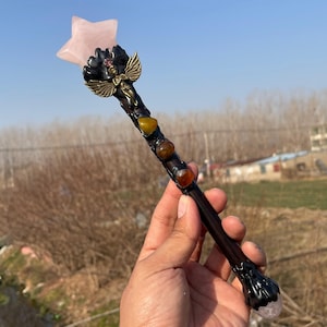 May include: A handcrafted wand with a pink star-shaped crystal at the top. The wand has a dark brown handle adorned with gemstones and a decorative angel figure. The wand is held in a hand against a natural outdoor backdrop.