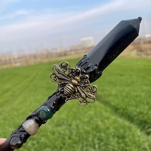 Op de afbeelding: Zwarte toverstaf met een zwarte kristallen punt, een gouden bij-bedel en groene en witte stenen.