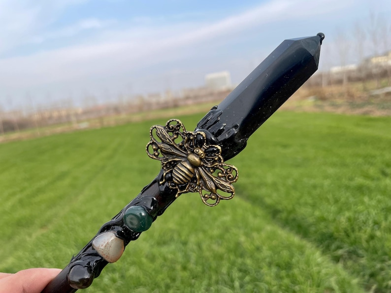Op de afbeelding: Zwarte toverstaf met een zwarte kristallen punt, een gouden bij-bedel en groene en witte stenen.