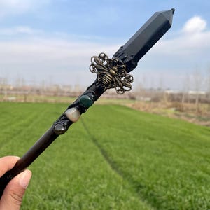 Op de afbeelding: Een zwarte kristallen toverstaf met een bij-bedel en groene en witte stenen. De toverstaf wordt in een hand gehouden tegen een groene grasachtige achtergrond.