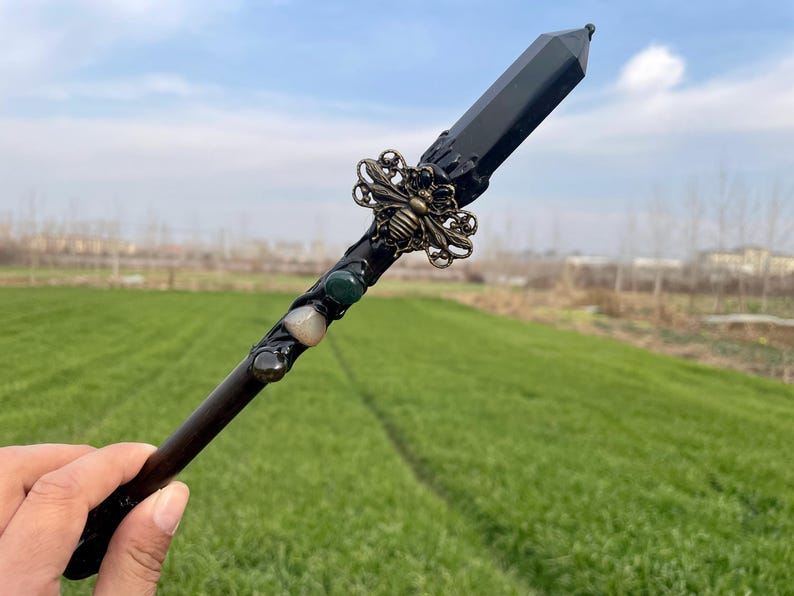 Op de afbeelding: Een zwarte kristallen toverstaf met een bij-bedel en groene en witte stenen. De toverstaf wordt in een hand gehouden tegen een groene grasachtige achtergrond.