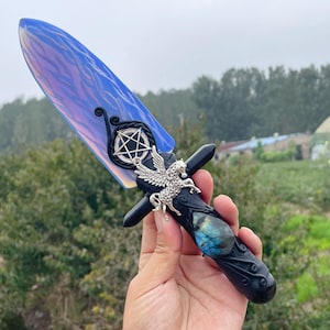 Puede incluir: Una daga decorativa con una hoja iridiscente azul y morada. El mango negro presenta detalles plateados, incluyendo un pentagrama, un caballo alado y una piedra preciosa azul. La daga se sostiene en una mano.