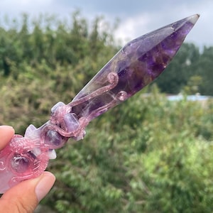 May include: A hand-held, decorative crystal dagger with a purple and pink color scheme. The blade is a deep purple, transitioning to a lighter hue at the tip. The handle features intricate carvings and a pinkish-purple hue, adding to its ornamental appeal.