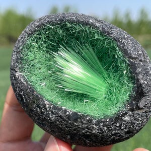 Puede incluir: Una geoda con un exterior gris oscuro y un interior hueco lleno de cristales verdes. Los cristales están dispuestos en un patrón radial, creando un efecto de cepillo.