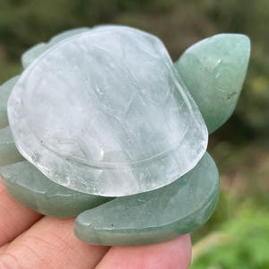 Könnte beinhalten: Eine geschnitzte Schildkrötenfigur aus durchscheinendem grünem und klarem Stein. Der Panzer der Schildkröte ist heller, fast weiß, während der Körper und der Kopf dunkler grün sind. Die Figur wird in einer Hand gehalten.