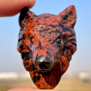 Puede incluir: Una figurita de cabeza de lobo tallada en jaspe rojo y negro. Los ojos del lobo son negros y el pelaje es rojo y negro moteado.