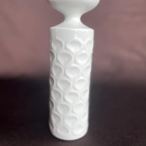 Puede incluir: Un jarrón de cerámica blanco con un patrón de gotas de agua repetido y texturizado. El jarrón tiene una parte superior acampanada en forma de pétalo y un cuerpo cilíndrico. El jarrón mide unos 20 cm de alto.